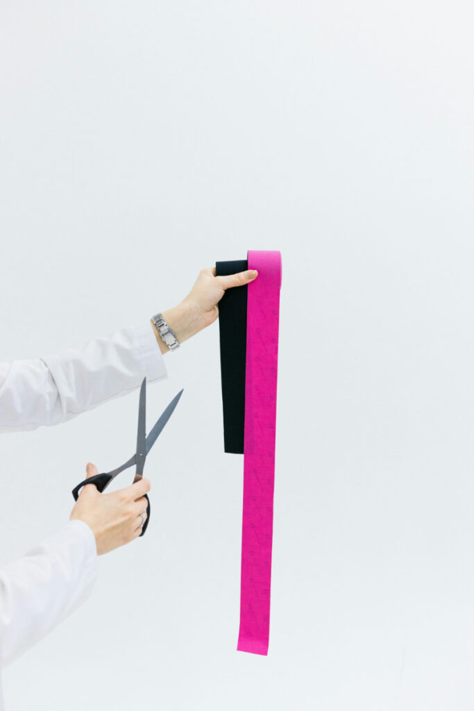 A person in a white coat cutting pink and black kinesiology tape with scissors, preparing it for therapeutic use.
